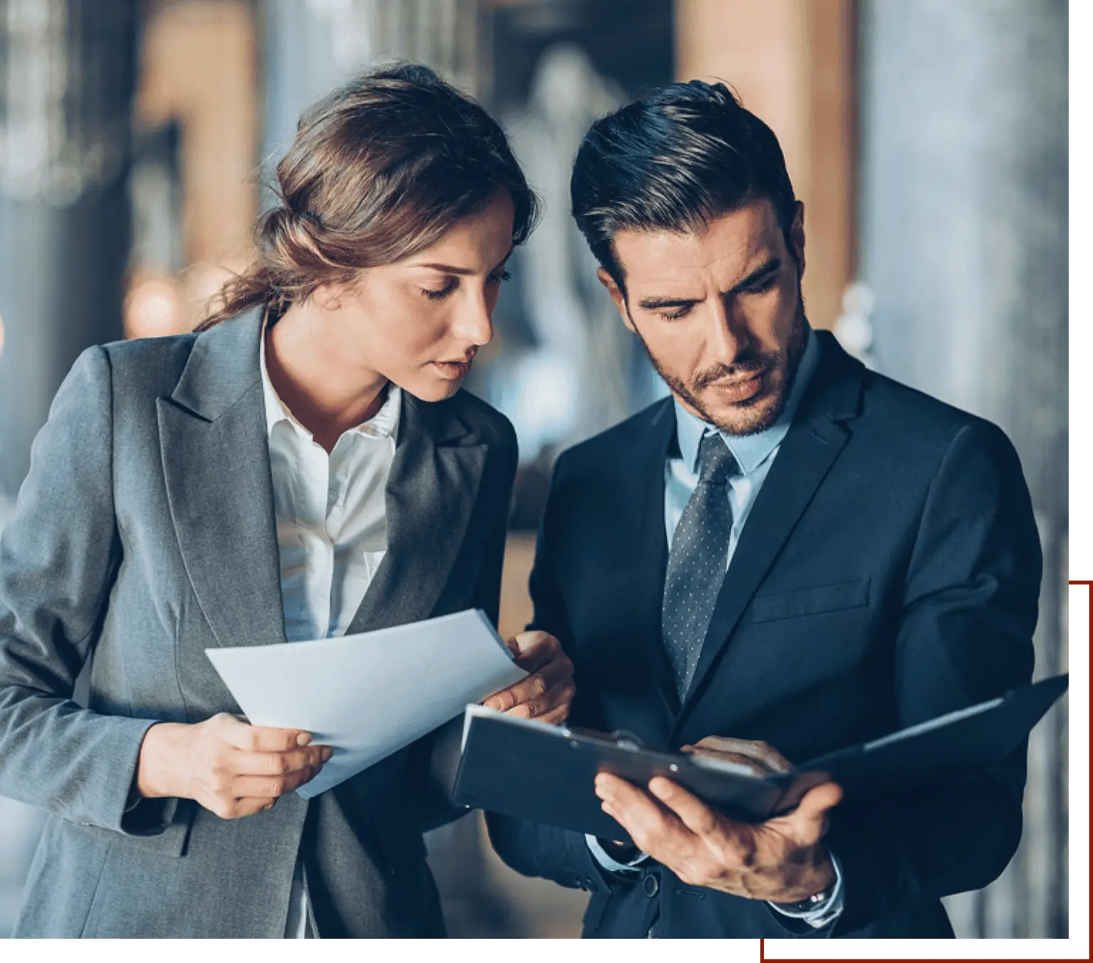 Man and woman in business attire looking at paperwork while standing in front of a blurred background