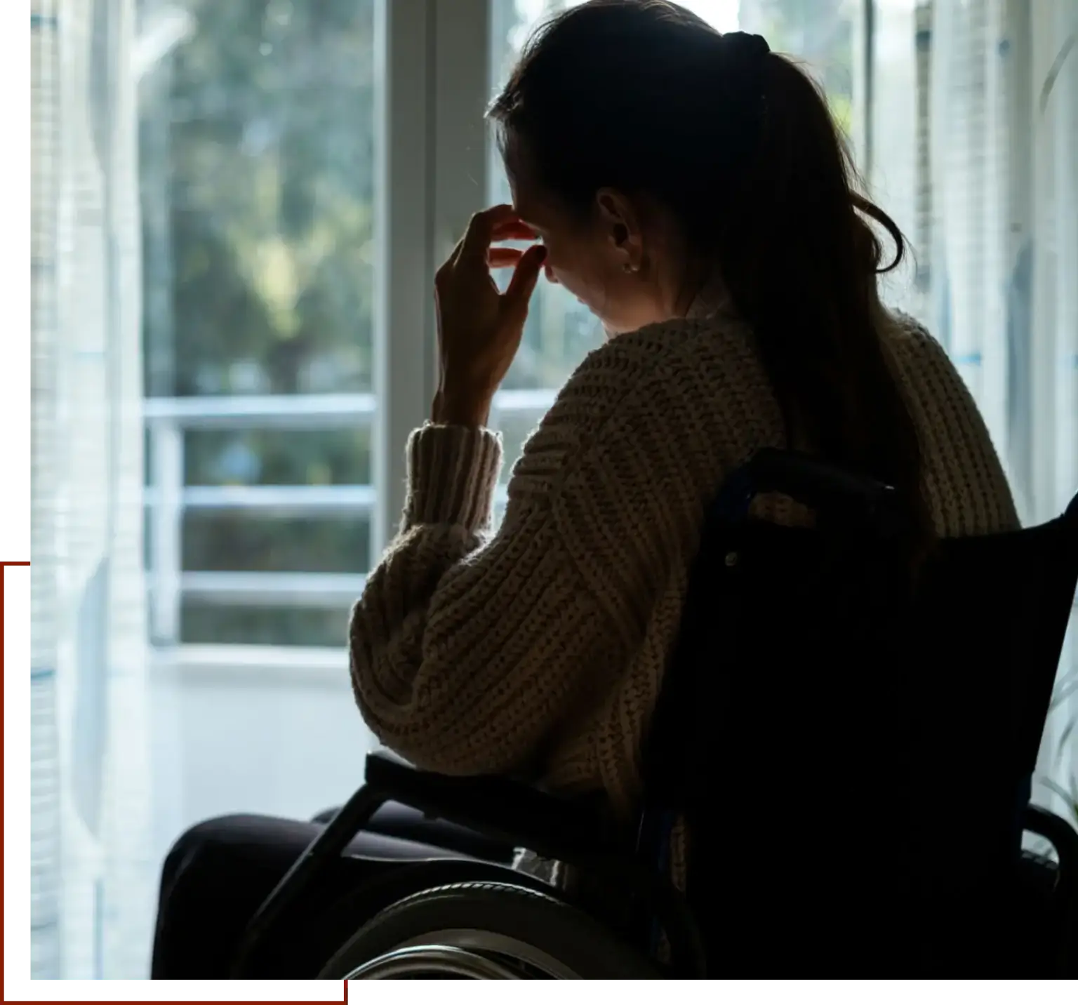 Woman in a wheelchair, wearing a sweater, looking out the window with a look of distress on her face